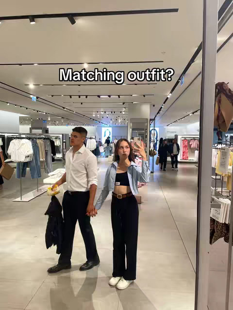 A couple poses in a clothing store. The male figure on the left wears a white shirt and black pants, holding a jacket and a snack. The female figure stands to the right, holding a phone and wearing a crop top, a blue shirt, and black pants.
