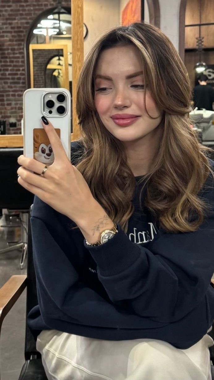 A young woman is taking a selfie in a salon. She has long, wavy brown hair with highlights and is wearing a navy blue sweatshirt, a gold watch, and a ring. She's holding a white phone with a cute case, and her facial expression is pleasant. The background appears to be a hair salon, with mirrors and styling equipment visible.