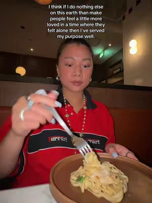A woman is shown seated at a restaurant table. She is wearing a red Ferrari polo shirt and holding a fork over a plate of pasta. Text overlay is added at the top of the picture.