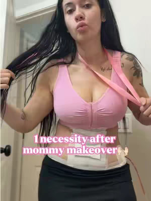 A woman with long black hair is posing, holding her hair with both hands. She is wearing a pink sports bra, and her torso shows post-surgery medical bandages. The background is a blurry room, likely a bathroom, with a door frame visible.