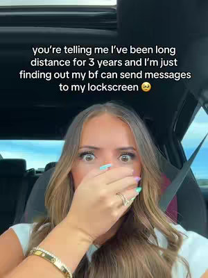 A woman sitting in a car with an expression of shock on her face, she is covering her mouth with her hand. The background is out of focus, showing the interior of a car.