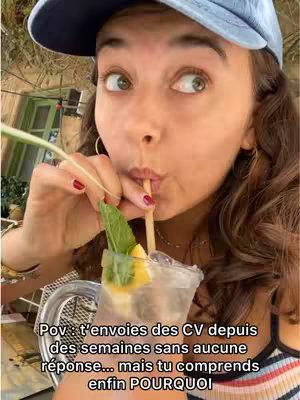 A young woman with dark hair and a blue cap is seen in close-up, looking upwards with a surprised expression while sipping from a straw; in front of the face a clear glass of a drink with ice and mint is visible.