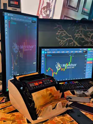 The image shows a desk setup with multiple computer monitors displaying financial charts, a money counter, and stacks of cash, suggesting a trading or finance-related theme. The scene is brightly lit, with the monitors as a primary focus and cash distributed around the desk.