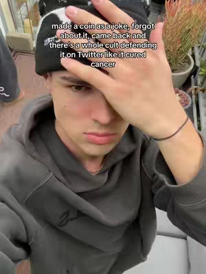 A young man wearing a black and white beanie and a gray hoodie covers part of his face with his right hand. The text overlay explains a joke about a coin that has now grown into a cult following.