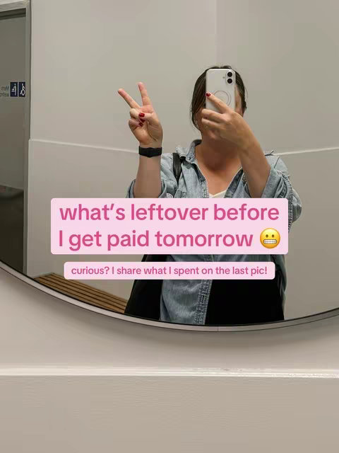 A person takes a mirror selfie in what appears to be a bathroom setting. The person is positioned in the center, slightly angled, holding a smartphone in front of their face, obscuring it from view. They are wearing a denim shirt and have their right hand raised, displaying a peace sign. In the center of the image, there's overlaid text in a pink rectangle that reads: 'what's leftover before I get paid tomorrow', followed by an emoji. Below this, there is smaller text: 'curious? I share what I spent on the last pic!' The background is a plain pale gray wall with a circular mirror, around which the selfie is framed, and part of the bathroom's accessories is visible.