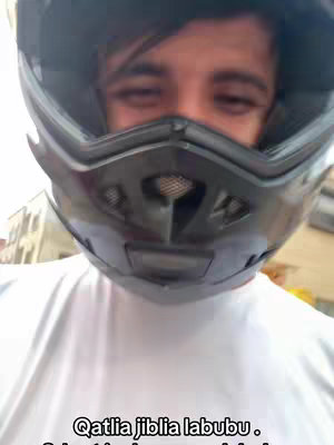 A selfie-style video cover image featuring a person wearing a black motorcycle helmet, obscuring the upper part of their face. The person is looking down at the camera with some visible facial features while wearing a white t-shirt. Text overlayed at the bottom of the image.