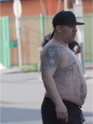 A man with tattoos on his chest and arm stands on a street. He wears a black cap and is bare-chested. Background includes a green fence and an orange building.