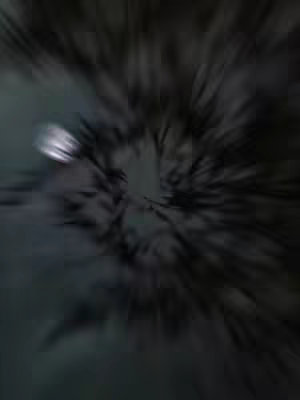 An abstract image featuring radial blur effects, creating a sense of movement and depth. The color palette is dark with a few areas of light, particularly in the upper left corner and center, where a blurred, bright spot appears.