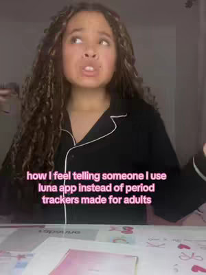 A young woman with curly hair is looking upwards with an animated expression. She's wearing black pajamas with white piping, sitting at a desk. The lower portion of the image  includes a desk with papers and a pink object on it. The image has a pink and neutral color palette.