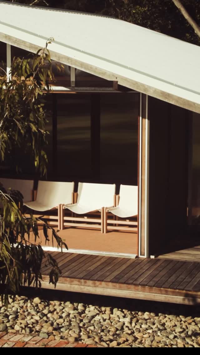 A minimalist, outdoor scene showcasing a modern architectural design. The focus is on three white chairs, with light wooden frames, elegantly arranged on a raised platform. The chairs are positioned inside a doorway that leads out onto a deck. The deck is bordered by pebbles below. A tree is on the left, with foliage framing the corner of the building.
