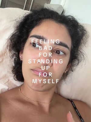 A selfie-style shot featuring a woman lying down in bed with text overlayed on her face. The text reads "FEELING BAD FOR STANDING UP FOR MYSELF". The woman has dark curly hair and is looking slightly off-camera with a thoughtful expression. She's wearing a thin gold necklace and a black top, suggesting a relaxed, intimate setting. The lighting is soft, and the background suggests a bedroom environment.