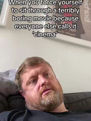 A medium shot of a man with a beard, looking at the camera with a somewhat bored or exasperated expression. The background includes a section of a wall with a framed picture, a poster, and a light-colored wall. The text overlay suggests a relatable experience of sitting through a boring movie.