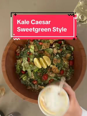 The cover image of a food-related video shows a wooden bowl filled with a salad. Within the bowl, a colorful mix of ingredients is visible, including green lettuce leaves, red cherry tomatoes, and what appears to be spiced roasted chicken and Parmesan crisps. A transparent glass with a light-colored liquid is positioned in the bottom right corner of the frame, the tip of the liquid pouring from a container into the bowl. The top of the original image features text on a rectangular red background that reads, 'Kale Caesar Sweetgreen Style.'