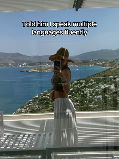 A woman is taking a selfie while standing on a balcony. She is holding the phone in front of her face. She wears a hat and long skirt. The background is a scenic beach landscape with mountains and blue water. In the background, there is lush greenery, and the sky is clear with some clouds. She is wearing a hat and holding a phone.