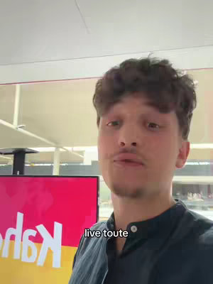A young man is filmed from a medium close-up, addressing the camera in an indoor setting. He has short, dark curly hair and a trimmed mustache and goatee. He wears a dark blue collared shirt. Behind him, a large screen displays the Kahoot! logo in red against a yellow background. To his right, a glimpse of an open space.