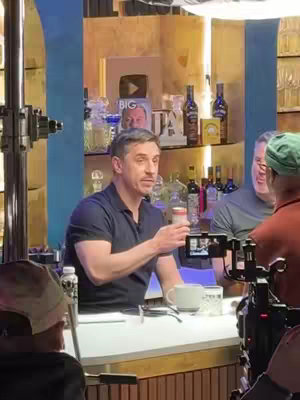 A man in a dark blue polo shirt holds up a vitamin can while speaking, in the background are two other people, bar and shelves with bottles.