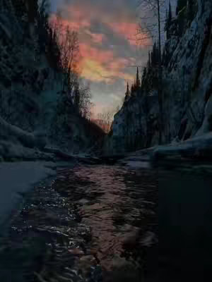 A serene landscape photograph captures a deep canyon or ravine with snow-covered sides, a river flowing through the center, and a vibrant sunset in the sky.