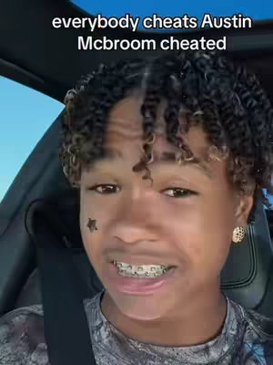 A young man with curly hair, a star-shaped face tattoo, and braces smiles in a car. Text overlayed on the image reads: 'everybody cheats Austin Mcbroom cheated'. The background is a blurry view outside the car, likely during the day.