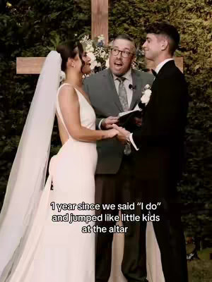 The cover image is a vertical shot taken at a wedding ceremony. The focus is on a couple holding hands as they stand before a cross. The background has green foliage. A man to the right, in a black suit and white flower in his lapel, smiles. A woman to the left wears a white dress and veil. Standing between them, an older man in glasses appears to be officiating the wedding, holding a book and speaking. The overall composition is brightly lit, with warm lighting that emphasizes the significance of the moment.