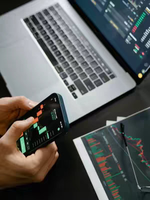 The image shows a person's hands holding a smartphone displaying a stock chart with red and green candlesticks. A laptop with a similar chart on the screen is visible in the background, along with financial documents and a pen. The scene is set on a dark surface. The image suggests a focus on investment and financial analysis.