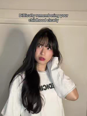 A woman with long black hair and bangs is looking upwards, with a slightly concerned expression. She is wearing a white t-shirt with black text on it. The background is a plain wall.