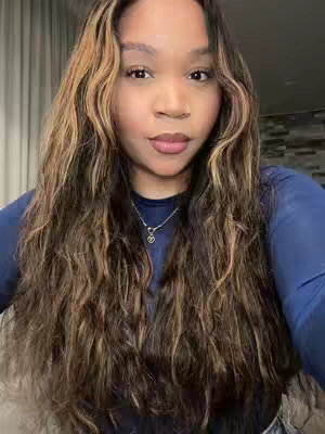 A medium shot of a woman with long, wavy brown hair with blonde highlights. She has a fair complexion, brown eyes, and a small nose ring.. She wears a dark blue long-sleeve top and a silver necklace with a heart pendant. The background consists of a blurry, neutral-toned setting with what appears to be a curtain