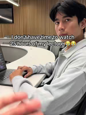 A young man in a light gray jacket is seated at a curved white desk in a lecture hall setting. He is looking towards the camera, his expression appearing thoughtful. Text on the image reads: 'I don't have time to watch 25 hours of my lecture'. The background includes wood paneling and a dark screen.