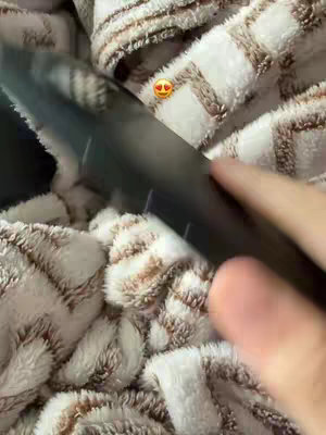 The cover image shows a close-up view of a person's hand and part of a black object, possibly a smartphone or a tool, resting on a fluffy, patterned blanket. The blanket has a beige and brown color scheme, providing a warm, textured background. In the frame, there is an emoji with heart eyes near the object. The shot is well-lit, with the focus on the texture and the interaction with the object.