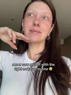 A woman with fair skin is captured from the chest up, centered in the frame. Her dark hair is styled down, and she is wearing a white t-shirt. She gazes slightly upward and to her right, with a thoughtful expression. Her left hand rests near her chin, her fingers gently touching her cheek. She wears hoop earrings and a ring. Overlayed text at the image bottom reads 'never saw myself with the right makeup routine' followed by a smiling emoji. The background is softly lit, suggesting an indoor setting.