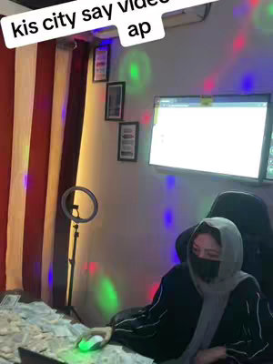 A woman wearing a hijab and face covering sits in a room strewn with money, in front of a computer. A ring light and framed pictures are visible on the wall. The video shows a woman sitting with a mouse in her hand in a lavish setting.