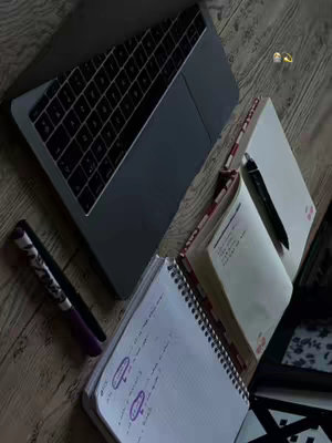 The image shows a workspace with a laptop, notebooks, and pens, all arranged on a wooden surface. The laptop, closed and positioned at an angle, dominates the top left. Several open notebooks are scattered, with handwritten notes visible in some. Pens are resting on or near the notebooks. The overall composition suggests a study or work environment, bathed in soft lighting with a warm color palette.