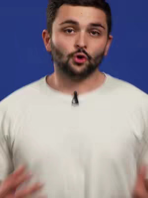 A man with a beard and short brown hair stands in front of a blue background, looking towards the camera with his mouth open. He's wearing a white t-shirt. The composition is a mid-shot that focuses on his face and torso.