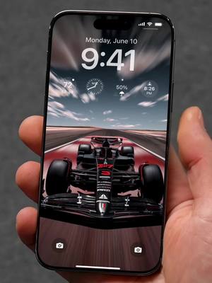 A hand holding a black iPhone displaying a close-up of a Formula 1 race car on a track. The phone's screen also shows the time and weather information, suggesting it's the lock screen. The background is a blurred image of the racetrack under a partly cloudy sky.