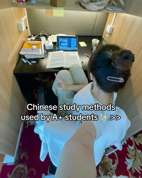 A selfie-style shot of a woman studying at a desk. She's seated with her back to the camera, positioned within a study cubicle. The desk is cluttered with books, an open tablet displaying a screen, and various other study materials. The woman is gesturing a 'peace' sign with her hand. The text overlayed on the image reads 'Chinese study methods used by A+ students'. The background features a carpeted floor and study area, contributing to the impression of an inside studying environment.