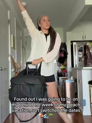 A woman stands in a room with her arm raised, holding a black suitcase. She is wearing a shirt over a black top and shorts, with a headband. The background shows a kitchen with a refrigerator and various other items.