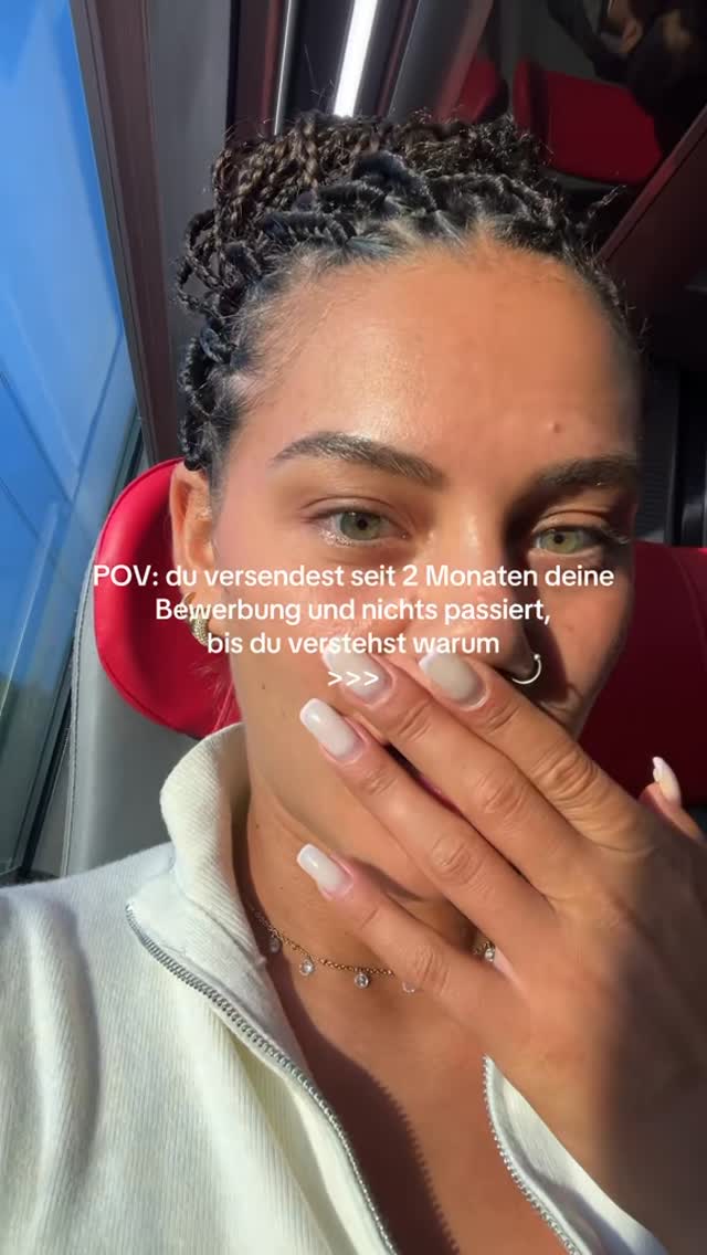 A close-up shot shows a fair-skinned person with long, dark hair styled in small braids. They are in a train, with a red seat visible behind them. They show a part of their face from their eye to their mouth, and their hand covers the lower half of their face with a long square nail manicure. The text overlay reads, "POV: du versendest seit 2 Monaten deine Bewerbung und nichts passiert, bis du verstehst warum >>>".