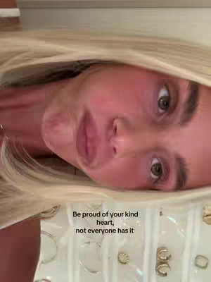 A close-up shot of a woman's face angled slightly to the side. Her blonde hair frames her face. The text overlay reads, 'Be proud of your kind heart, not everyone has it'. Behind her is a partial view of what seems to be a jewelry display. The overall tone is positive and encouraging.
