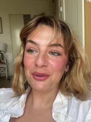 A selfie-style shot featuring a woman indoors. The woman has light skin, blonde hair with bangs, and visible makeup including blush. She appears to be speaking or reacting to something off-camera. The background shows a partially visible door and wall, with soft natural lighting.