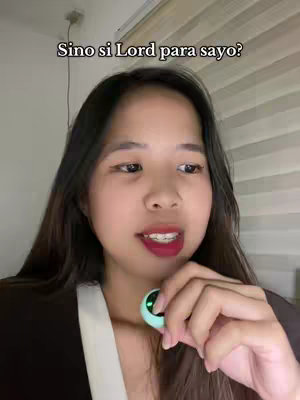 A young woman with long black hair is centered in the frame, looking at the camera and speaking. She holds a small, round, light-green device and is wearing a black top. The background is blurred, suggesting an interior setting with a window or blinds visible. The text overlay, "Sino si Lord para sayo?" (Who is the Lord to you?), runs along the top, in white font against a dark background.