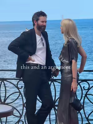 A man and a woman stand close to each other on a balcony overlooking a body of water, the background blurred. The man, on the left, wears a black blazer over a white shirt and black pants. The woman on the right, in a metallic silver dress, slightly turns towards him. Text overlayed includes the phrase 'this and an argument'.