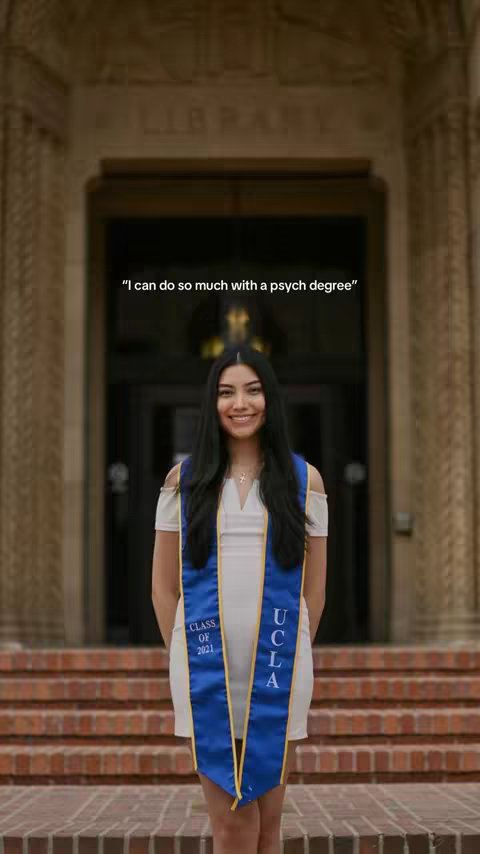 A young woman with long black hair smiles confidently at the camera, wearing a white dress and a blue and gold UCLA graduation sash. She stands in front of a large brick building with arched doorways and 'LIBRARY' etched above, suggesting a university setting. The photo is well-lit, emphasizing her positive expression.