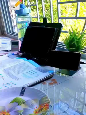 The image shows a study setup on a windowsill. There's an open textbook, a notebook, a phone holder with two phones, a water bottle, a small clock, and a plate. The background includes a view through a window, with greenery visible outside. A pencil with a drawing is added on top.
