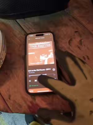 A phone displaying the album art and a song title by "The Smiths" is held against a wooden surface. A hand with potentially partially painted fingers is partly visible. The album art is in warm tones.