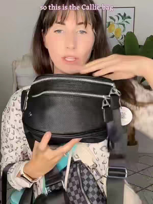 A woman holds up a black, leather messenger bag to the camera, showcasing its features. The background is a blurred room with hints of decor.