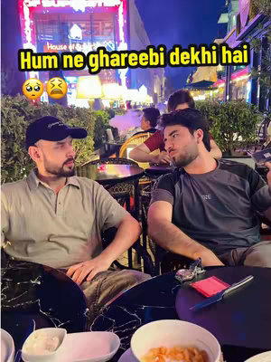 Two men are seated at an outdoor table, having a discussion. A text overlay at the top of the image reads a Hindi phrase. The scene is set in a restaurant or cafe, with blurred background elements suggesting city lights and other patrons.