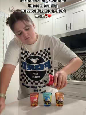 A person holding a beverage of some kind in their right hand, pouring it into three small shot glasses that have Sonic characters on them. Behind them is a kitchen with white cabinets.