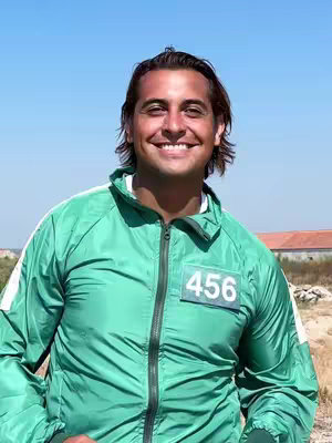 A smiling man with dark hair wearing a green tracksuit. The background shows a blue sky and outdoor area.