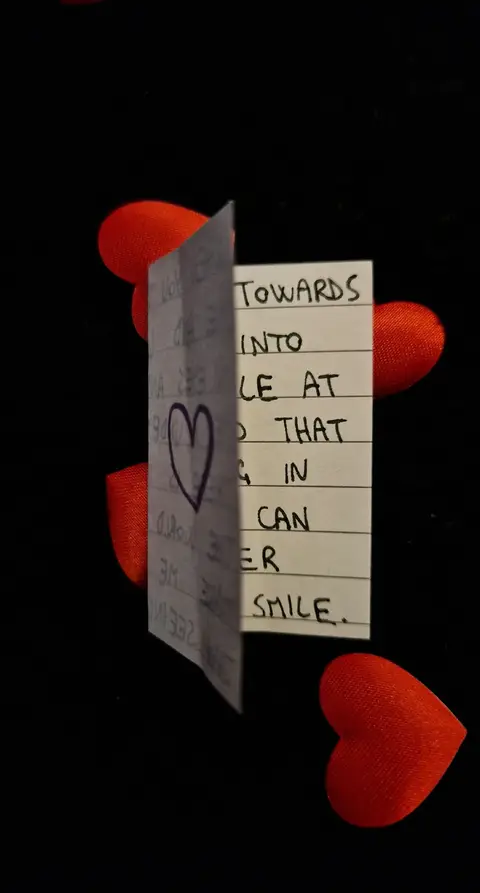 The image shows a black background with red heart decorations. A white folded card with handwriting is in the center of the image. The exposed side of the card has a large heart drawn in the middle. The text is written on the card as well. The red hearts around are placed randomly.