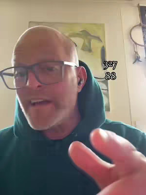 A bald man with glasses and a green hoodie is speaking with his right hand raised, with what appears to be art or wall decor in the background, and text overlay with numbers.