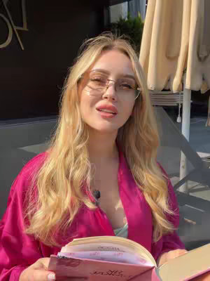 A woman with blonde hair wearing glasses and a pink top is sitting outside. She is holding a book and looking up, possibly speaking. An umbrella is visible to the right of the frame.